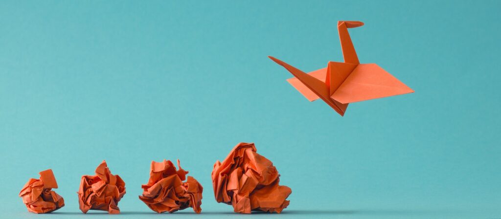 Five orange paper balls sit on a surface; one transforms into an origami crane that appears to be flying upwards against a blue background.