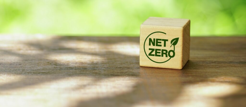 A wooden block with a green NET ZERO logo and a leaf symbol sits on a sunlit wooden surface, with a blurred natural background.