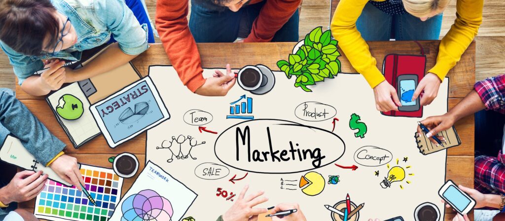 A group of people sit around a table with marketing-related drawings, charts, and notes. Hands hold coffee cups, a color palette, a tablet, and notebooks. The word Marketing is written in the center with diagrams and icons around it.