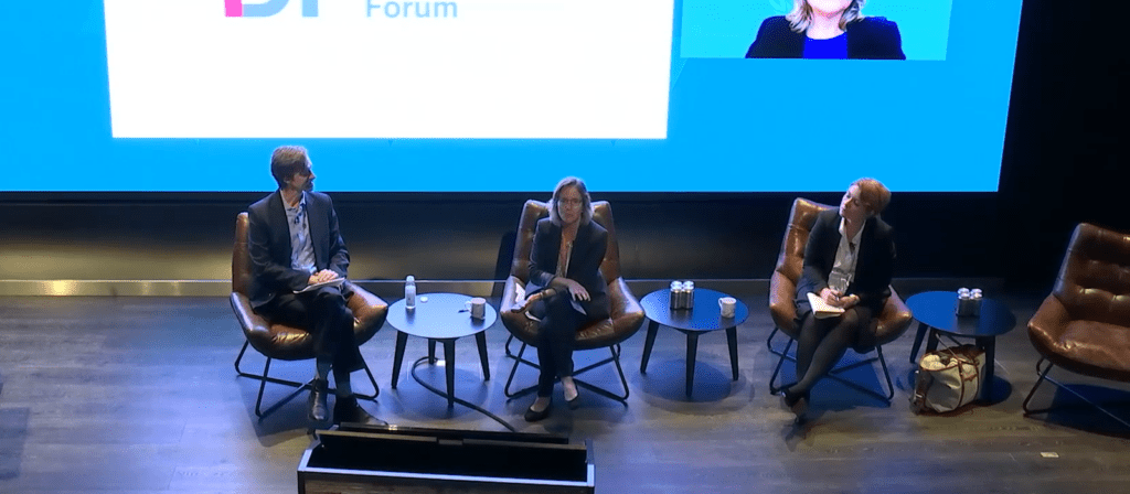 Three people sit on a stage in leather chairs, participating in a panel discussion. Behind them is a large screen showing part of an event logo and a speaker’s video feed. Small tables hold water bottles.