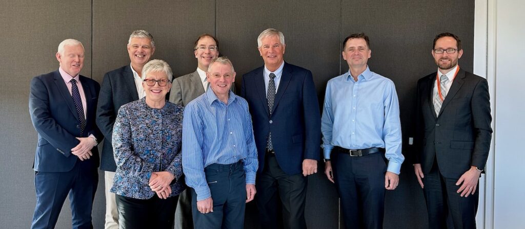 Eight adults, six men and two women, stand side by side indoors against a plain wall, dressed in business and smart-casual clothing, smiling at the camera.