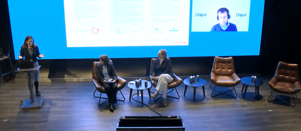 A woman speaks at a podium on stage next to two seated panelists and three empty chairs, with a large screen behind them displaying a video call participant and presentation slide.