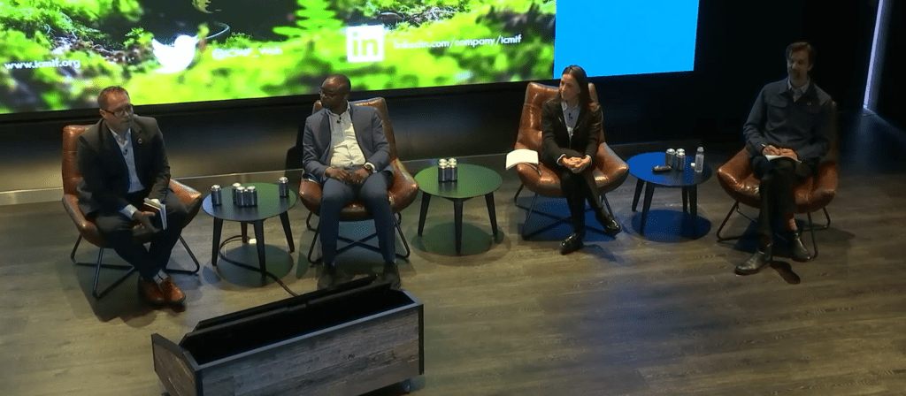 Four people sit in chairs on a stage, spaced apart, participating in a panel discussion. Three small tables with water bottles are between them. A digital display with social media handles is visible behind them.