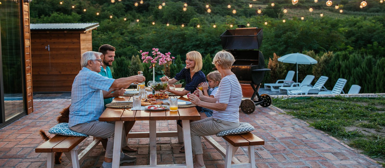 Five people of varying ages sit around an outdoor picnic table, enjoying a meal together on a patio. String lights hang above, and lounge chairs and greenery are visible in the background.