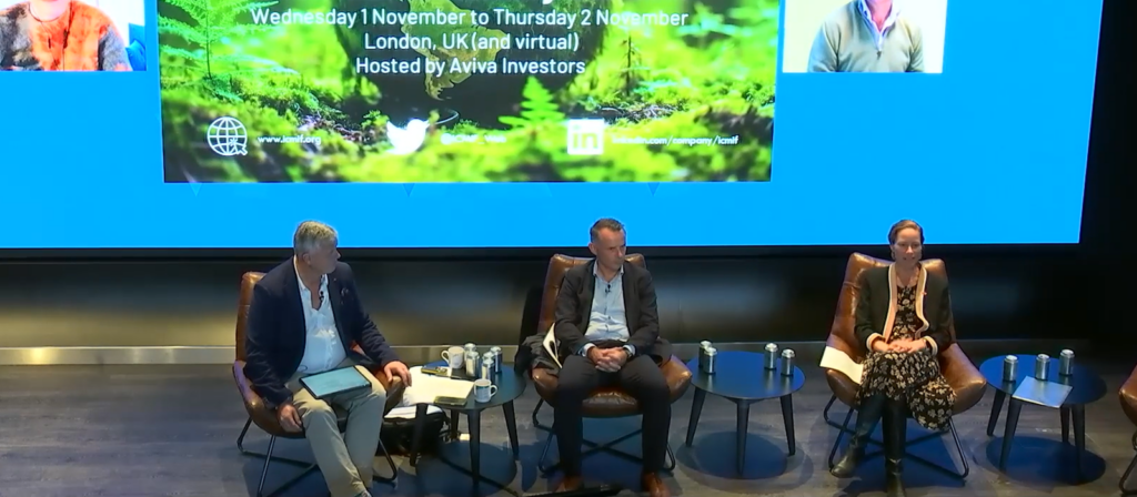 Three panelists sit on stage with microphones during a conference. Behind them is a large screen displaying event details, including dates, location (London, UK), and the host, Aviva Investors.