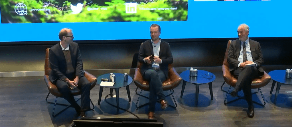 Three men sit in brown chairs on a stage, participating in a panel discussion. Two small tables with water bottles are between them, and a large blue screen with social media logos is displayed behind them.