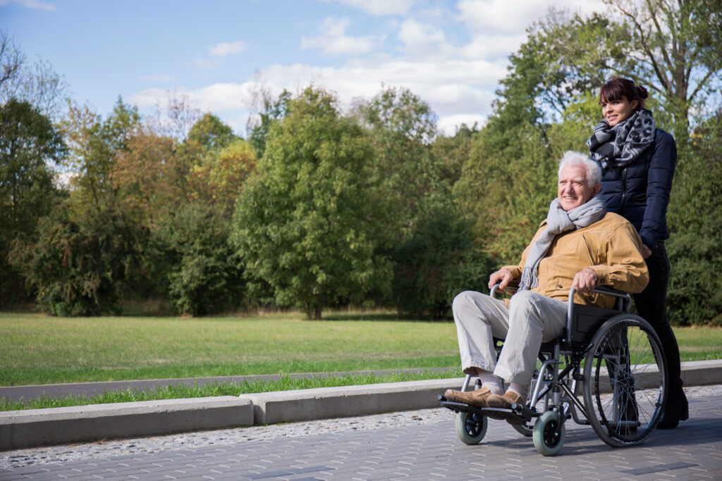 An older man in a wheelchair is being pushed by a woman outdoors on a paved path, surrounded by green trees and grass, both dressed warmly and smiling.