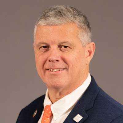A middle-aged man with short gray hair, wearing a dark suit jacket, white dress shirt, and an orange polka dot tie, smiles against a plain gray background.