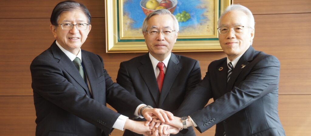 Three men in suits stand together indoors, each placing one hand over the others’ in a gesture of unity. They are smiling slightly, with a framed painting of fruit on the wall behind them.