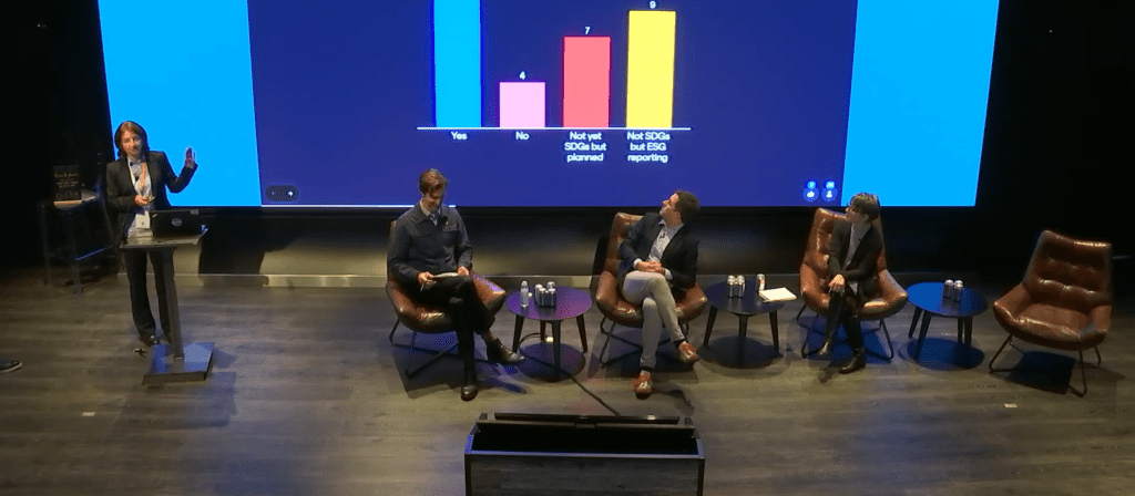 A person stands at a podium speaking while three others sit on stage. Behind them, a screen displays a bar chart with four colored bars labeled: “Yes,” “No,” “Not yet, but planned,” and “Not SOX but ESG reporting.”.
