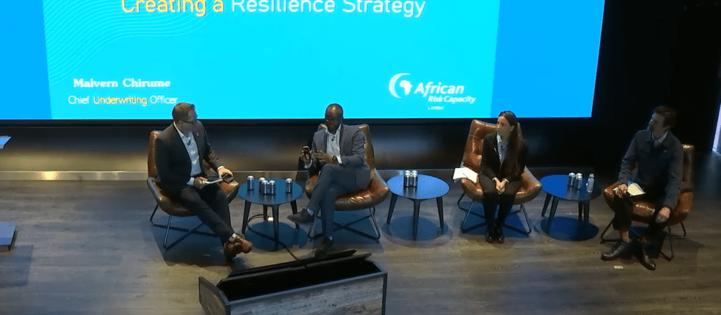 Four panelists sit on stage in armchairs, engaged in discussion under a large screen displaying “Creating a Resilience Strategy” and “African Risk Capacity Limited.” Bottled water sits on tables beside them.