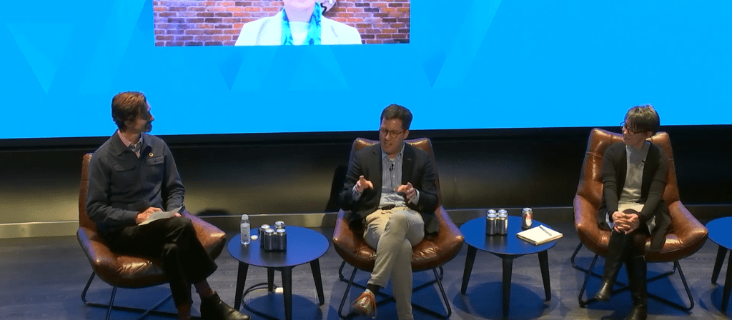 Three people sit in armchairs on stage, engaged in a panel discussion. Two small tables with water bottles and notebooks are between them. A large blue screen with a virtual participant is visible in the background.