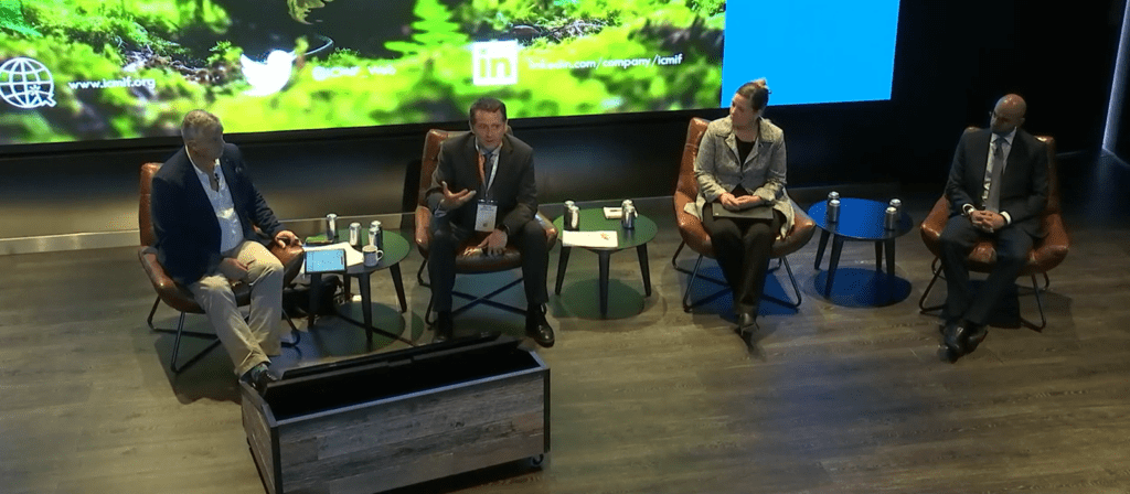 Four people in business attire sit on stage in armchairs for a panel discussion. A green and blue screen with social media icons is behind them. Tables with drinks are next to each chair.