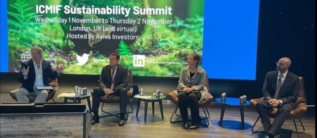Four panelists sit on stage during the ICMIF Sustainability Summit, with a presentation screen behind them displaying event details and social media links. The atmosphere appears professional and focused.