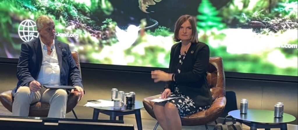 Two people sit on stage in armchairs with notepads, discussing in front of a green, nature-themed screen backdrop. Several cans and water bottles are on the tables between them.