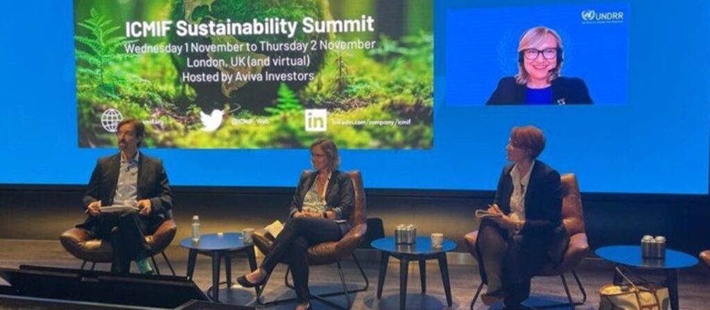 Three people sit on a stage panel at the ICMIF Sustainability Summit, with a large event screen behind them. A woman appears on the screen via video call. The setting is professional and conference-like.