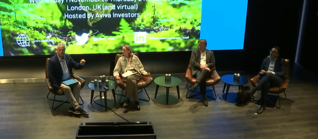 Four people sit on stage in armchairs for a panel discussion, with water bottles on small tables beside them. A large screen behind displays event details, mentioning London, UK, and host Aviva Investors.