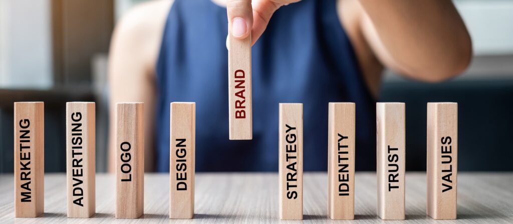 A person arranges wooden blocks labeled with “MARKETING,” “ADVERTISING,” “LOGO,” “DESIGN,” “STRATEGY,” “IDENTITY,” “TRUST,” and “VALUES,” while holding up a central block labeled “BRAND.”.