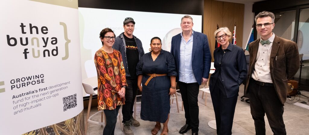 Six people stand smiling in a modern office space next to a large sign that says “the bunya fund.” Flags and a screen are visible in the background, suggesting a formal event or presentation.