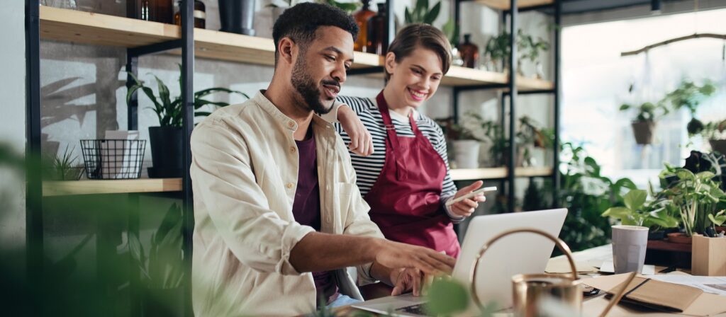 A man works on a laptop at a table surrounded by plants while a woman in an apron stands beside him, smiling and holding a smartphone. They appear to be collaborating in a bright, modern workspace.