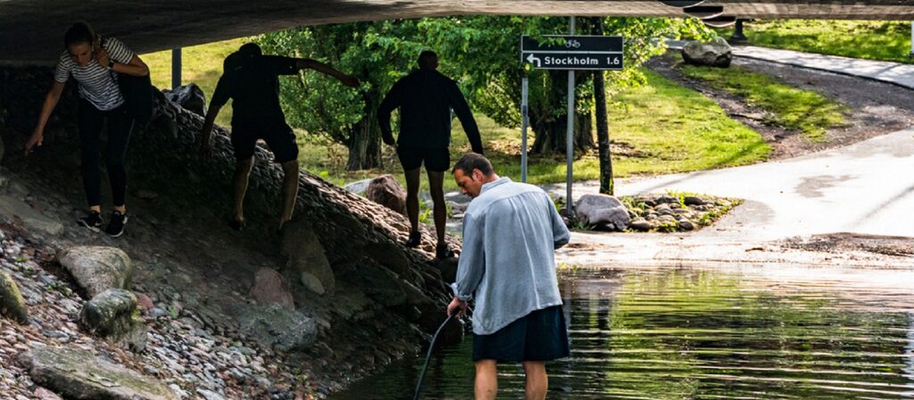 Four people walk under a bridge beside a shallow stream, with one person in the water and others navigating rocks. Trees and a sign pointing to Stockholm are visible in the background.