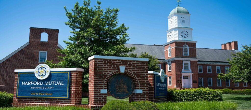 A red brick building with a white clock tower stands behind a large sign reading Harford Mutual Insurance Group on a sunny day, surrounded by green trees and neatly trimmed bushes.