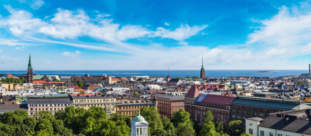 Panoramic view of a city with colorful buildings, church spires, and lush green trees in the foreground, under a bright blue sky with scattered white clouds, and the sea visible in the distance.