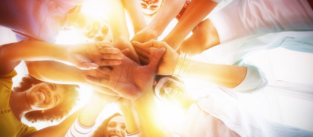 A diverse group of people stand in a circle, smiling and stacking their hands together in the center, viewed from below with sunlight streaming through their arms.