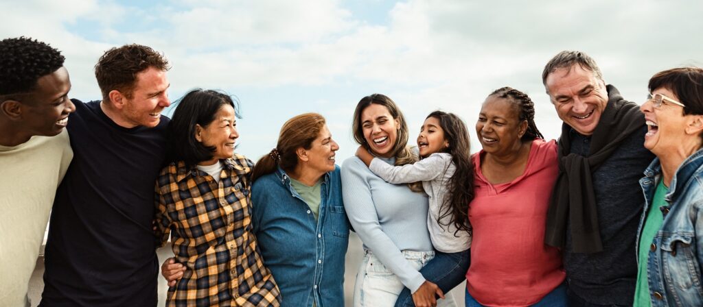 A diverse group of people standing in a row outdoors, smiling and laughing together. A young girl is being held and hugged by a woman in the center. The group appears happy and connected under a partly cloudy sky.