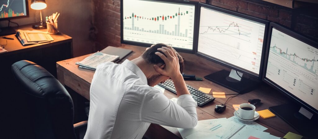 A stressed person in a white shirt sits at a cluttered desk, holding their head, surrounded by four computer monitors displaying complex financial charts and graphs. Papers, sticky notes, and a cup of coffee are on the desk.