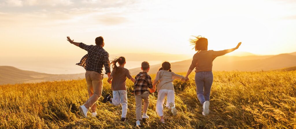A family of five runs joyfully through a grassy field at sunset, holding hands and spreading their arms, with rolling hills and a bright sky in the background.