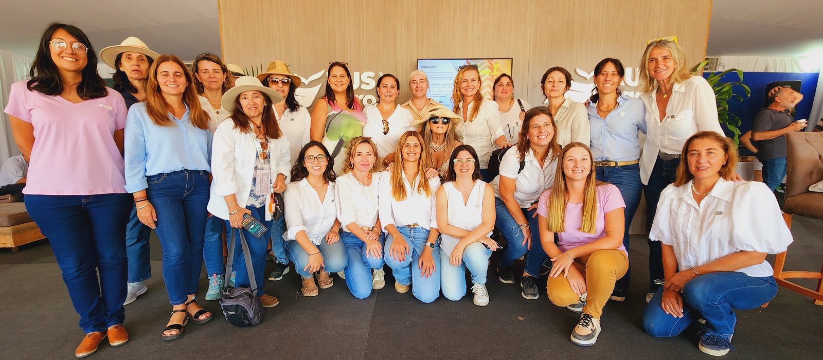 A group of women pose together indoors, smiling at the camera. Most are wearing casual outfits in light colors, such as jeans and white tops. The background features light wood paneling and a television screen.