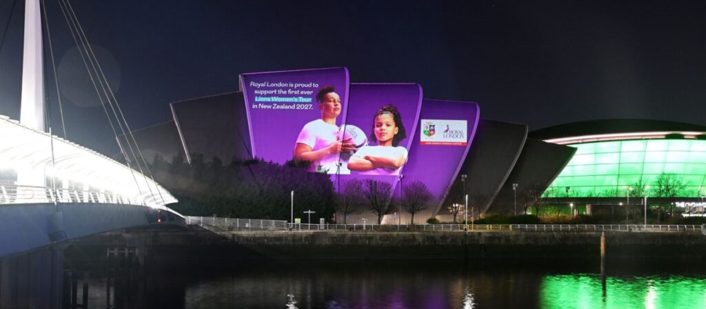 A building illuminated with purple lights displays a large projected image of two women in sports attire and text promoting Royal Londons support for the Womens British & Irish Lions in New Zealand 2027. A river and footbridge are in the foreground.