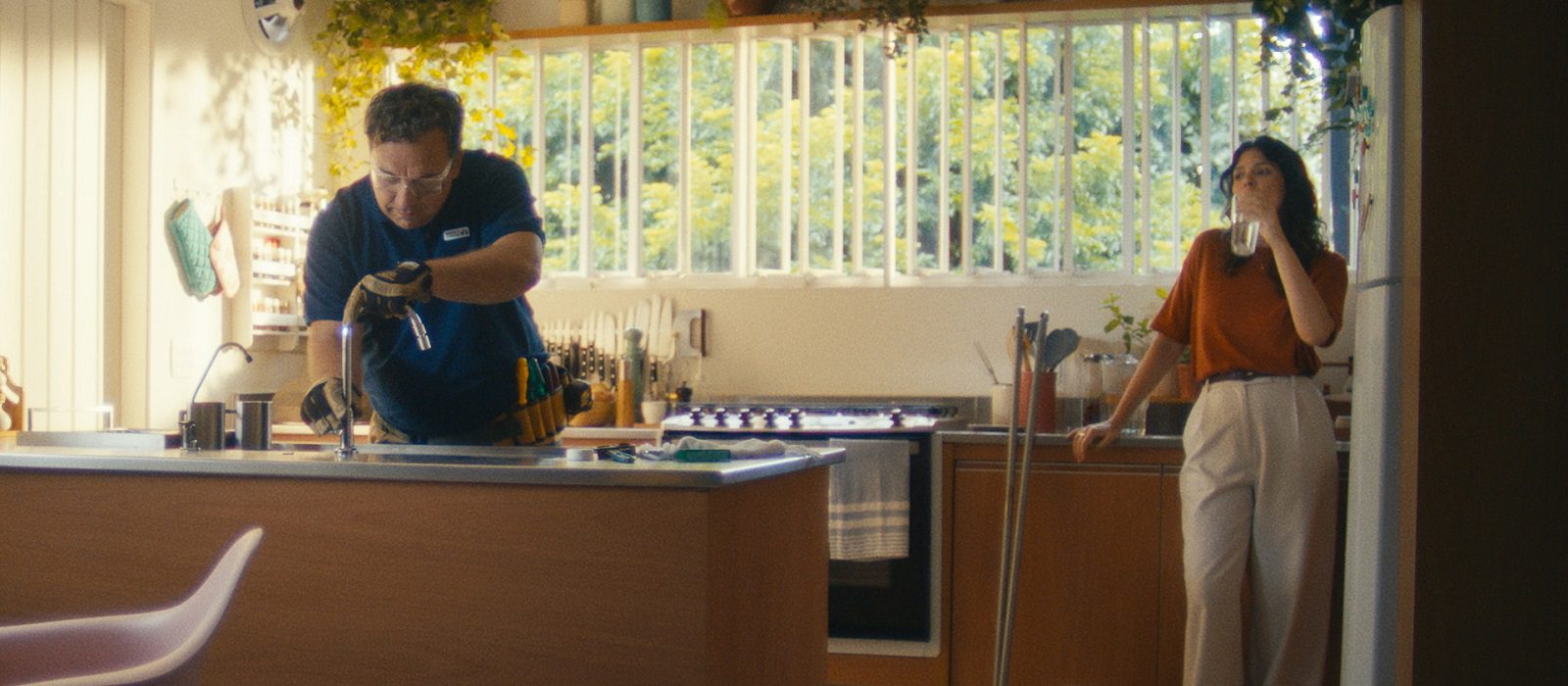 A man repairs a kitchen sink while a woman in an orange shirt stands nearby, drinking from a mug. The kitchen is bright with large windows and green plants on the shelves.