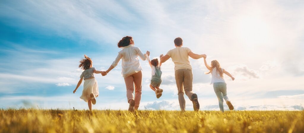 A family of five, viewed from behind, runs hand-in-hand through a sunny, grassy field under a blue sky with wispy clouds. The parents swing the youngest child between them as they all enjoy the outdoors together.