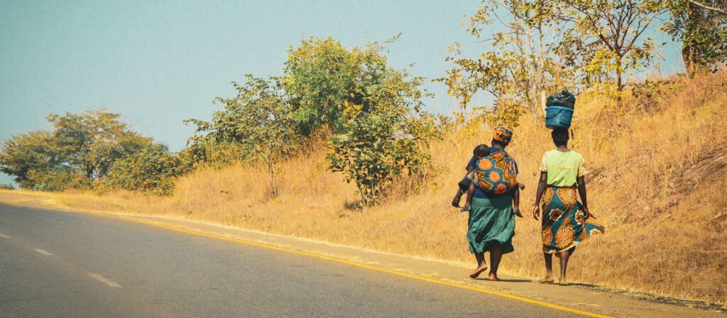 Two women walk along a rural road bordered by dry grass and trees; one carries a baby on her back, and the other balances a bundle on her head. Both wear colorful clothing and headscarves.