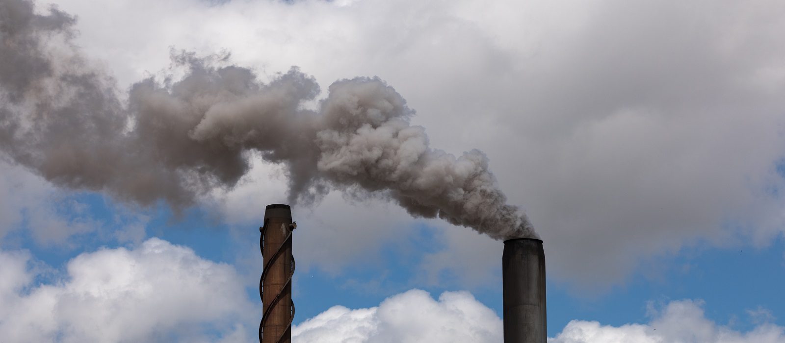 Two industrial smokestacks emit thick gray smoke into a partly cloudy sky, highlighting air pollution against a backdrop of white clouds and blue sky.