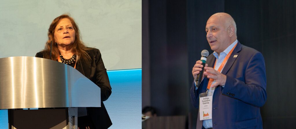 A woman stands at a podium speaking, while a man in a suit speaks into a microphone and gestures with his hand, both appearing to address an audience at a conference or event.
