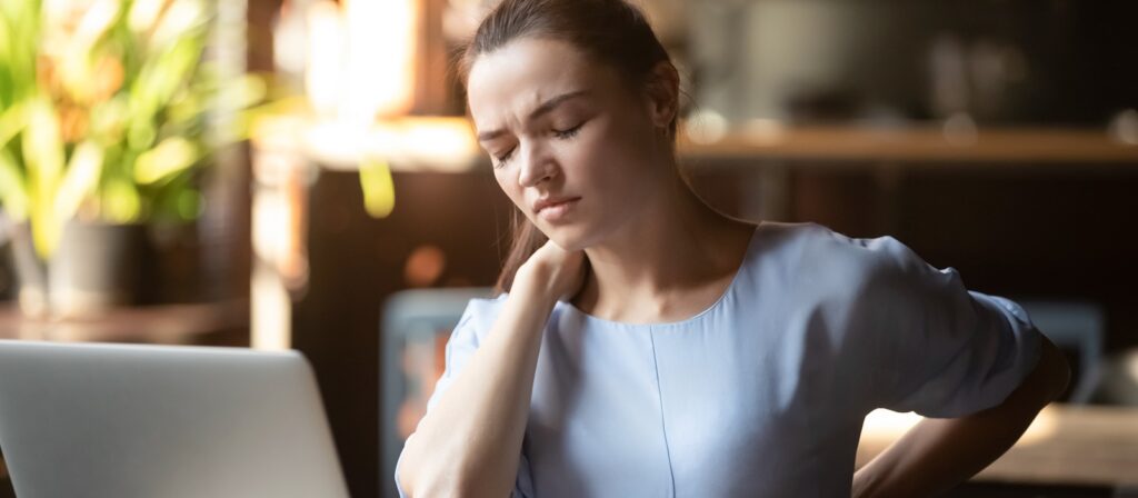 A woman in a light blue shirt sits in front of a laptop, eyes closed, and holds her neck and back in discomfort, possibly experiencing pain or tension. Bright indoor setting with plants in the background.