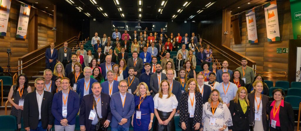 A large group of people pose for a photo in an auditorium, many wearing name badges and lanyards. The group is diverse in age and gender, standing and sitting on tiered seats, with conference banners visible on the walls.