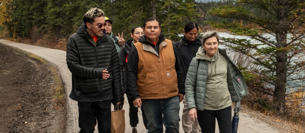 A group of people dressed warmly walk together along a riverside path bordered by trees, with one person holding a paper bag and another flashing a peace sign.