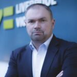 A man in a suit stands with arms crossed in front of a blue sign that reads LIVERPOOL and VIS with a green heart symbol and partially visible letters.
