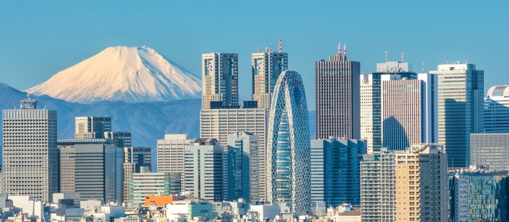 Tokyo skyline with modern skyscrapers in the foreground and Mount Fuji, snow-capped, rising in the background against a clear blue sky.