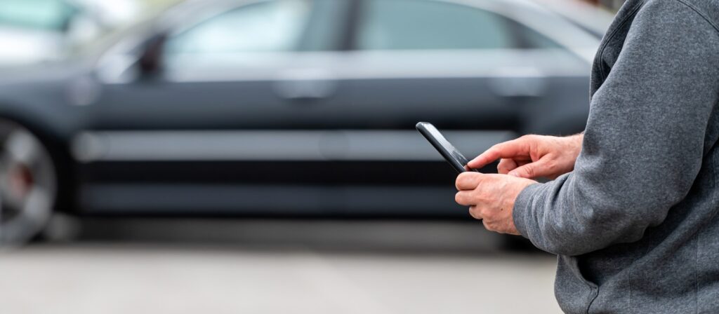A person wearing a gray sweater uses a smartphone outdoors, with a blurred black car parked in the background.