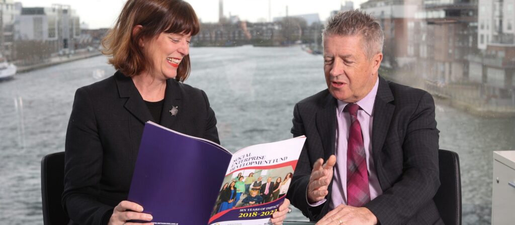Two people in business attire sit by a window overlooking a river, smiling and looking at a colorful document or report together. The document cover features group photos and text.