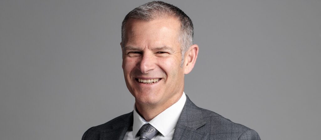A middle-aged man with short gray hair, wearing a gray suit, white shirt, and patterned tie, smiles warmly at the camera against a plain gray background.
