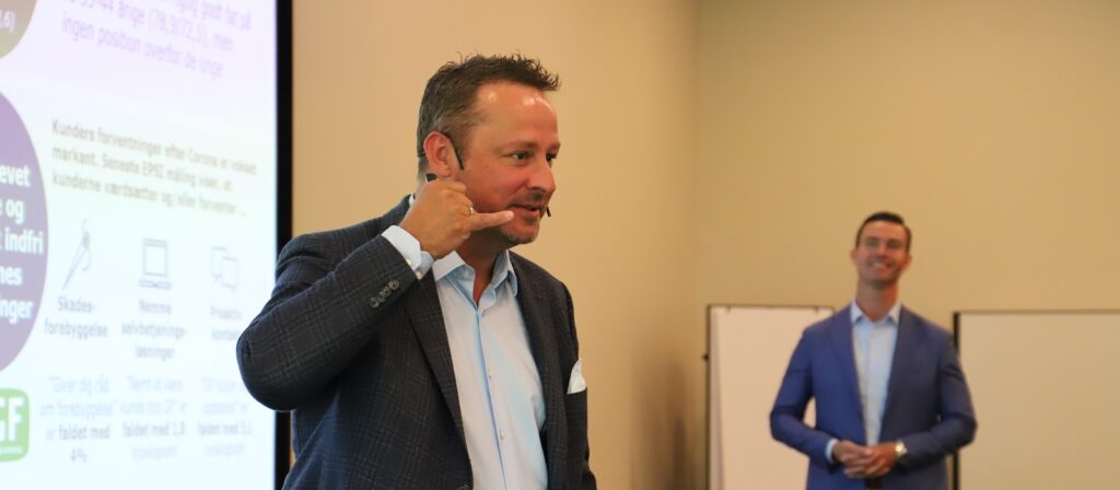 A man in a suit gestures with his hand as if using a phone while speaking in front of a projected presentation; another man in a suit stands smiling in the background.