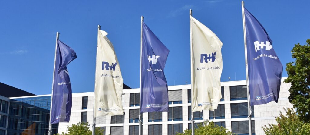 Five flags, three blue and two white, with the RH logo and German text wave in front of a modern building with large windows under a clear blue sky. Some green tree branches are visible on the right side.