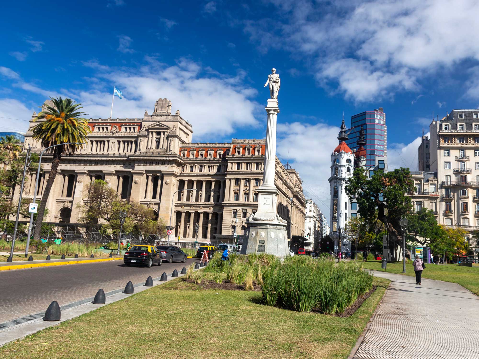 A sunny day in front of a historic courthouse building with columns, a tall white statue on a pedestal, palm trees, green grass, and a few people walking in Buenos Aires, Argentina.