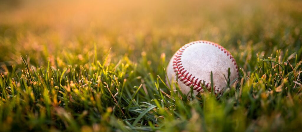 A baseball with red stitching rests on green grass, bathed in warm sunlight.